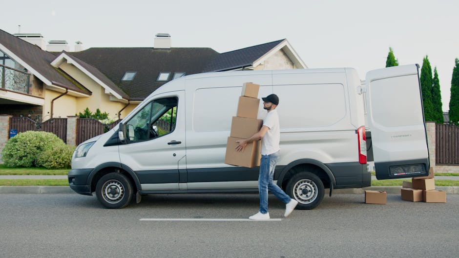A person wearing a dark jacket, black trousers, white sneakers, and a baseball cap is loading medium-sized, brown corrugated cardboard boxes into the back of a white delivery van. The boxes are sealed