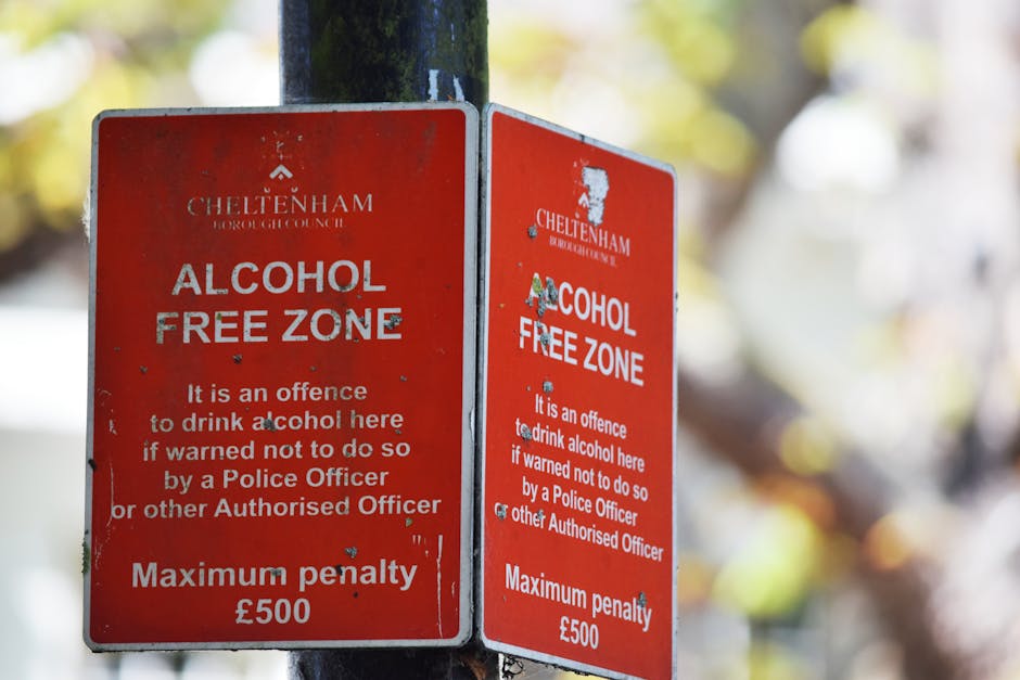 A pair of rectangular red metal signs are mounted on a dark green metal post, with one slightly overlapping the other. The signs feature white text and logos, indicating a designated 'Alcohol Free Zon
